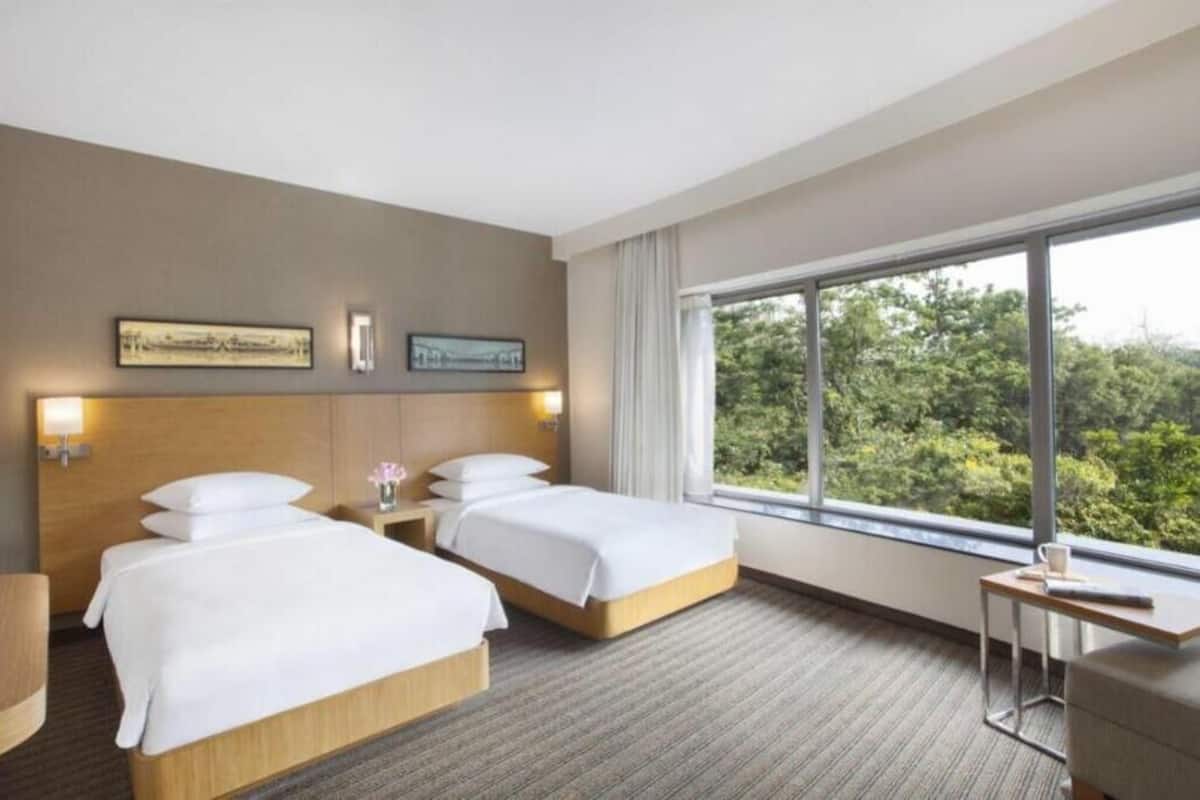 A hotel room with two beds, a small table, a nightstand, and a large window with a view of greenery.