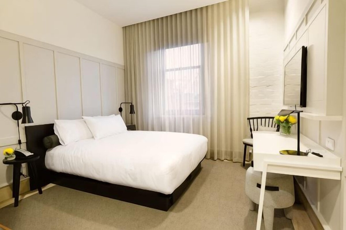 Una habitación de hotel con cama, mesitas de noche, escritorio, silla, espejo y ventana con cortinas.