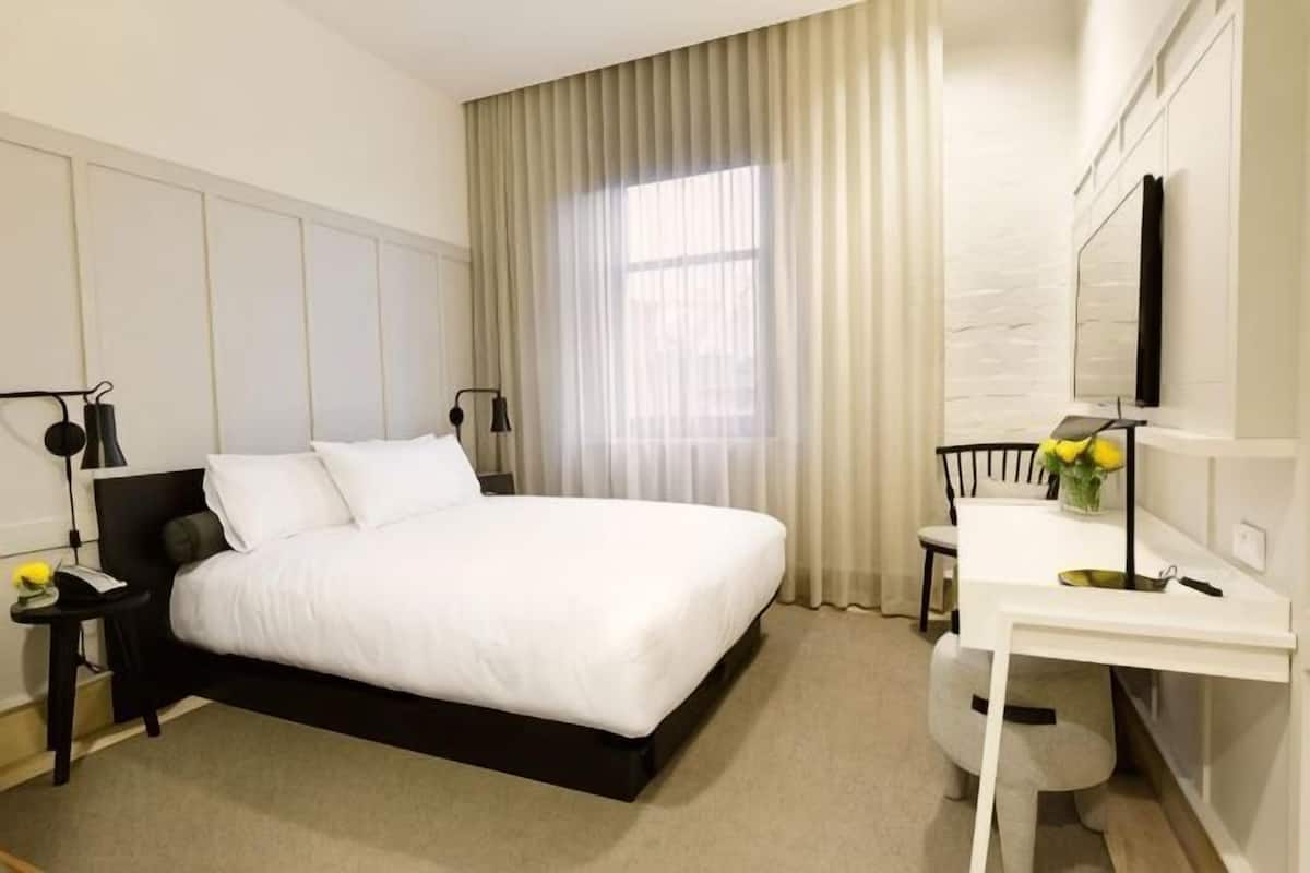 A hotel room with a bed, bedside tables, a desk, a chair, a mirror, and a window with curtains.