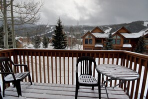 A wooden deck with two chairs and a table, overlooking a snowy mountain landscape with cabins.