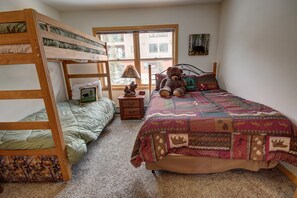 A room with bunk beds, a large bed with a colorful quilt, a teddy bear, a lamp, and a small table with a picture frame.