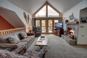 A cozy living room with a fireplace, a large TV, and a view of the outdoors through a large window.
