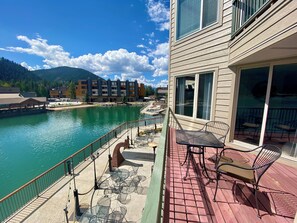 A balcony with outdoor seating overlooking a body of water, a building with large windows, and a view of a mountainous landscape in the background. - Lakeside 1481 by SummitCove Vacation Lodging (Keystone)