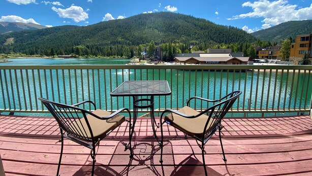 A deck with two chairs and a table overlooking a lake, with buildings and mountains in the background. - Lakeside 1481 by SummitCove Vacation Lodging (Keystone)
