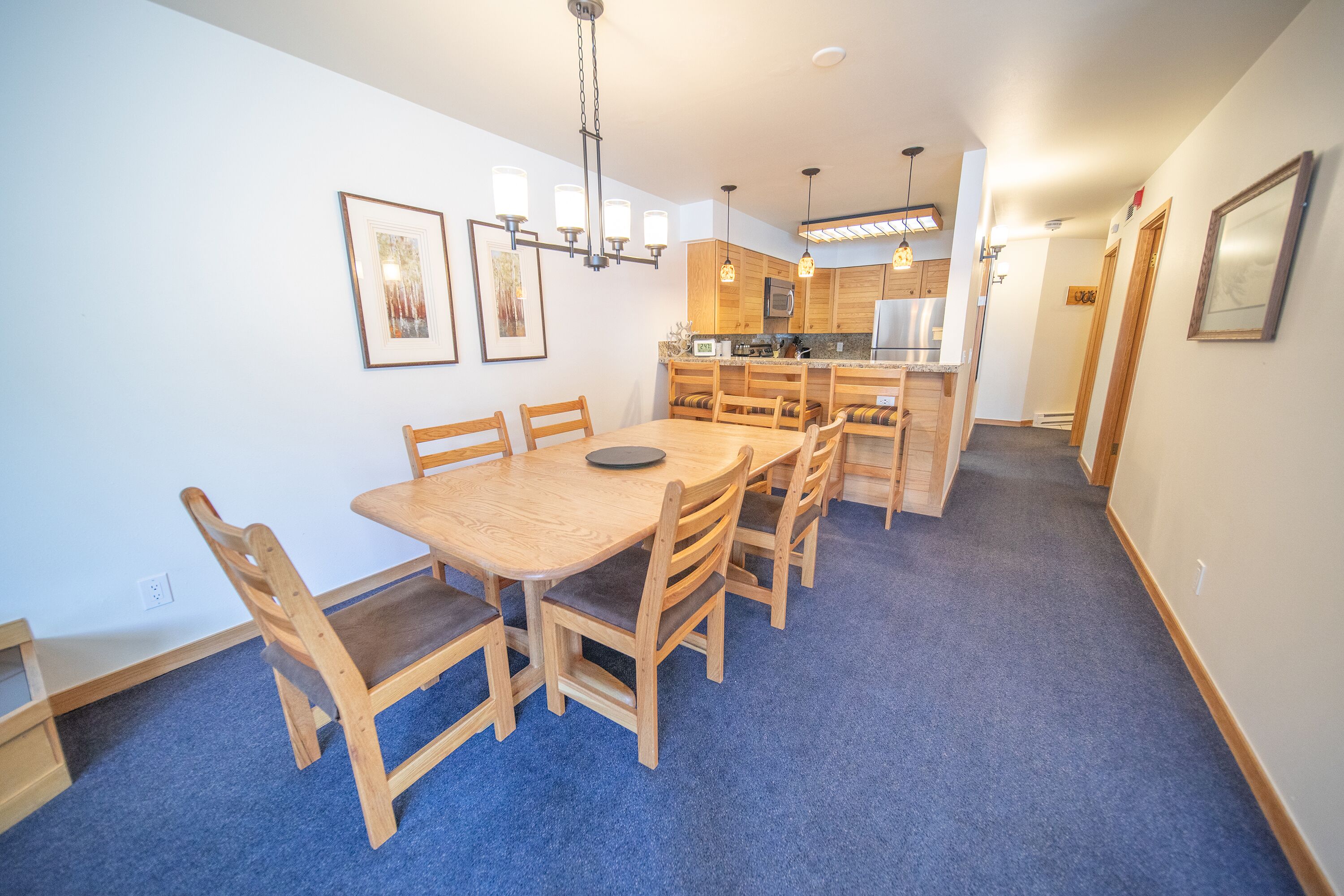 A dining area with a wooden table and chairs, a chandelier, and a kitchen in the background.