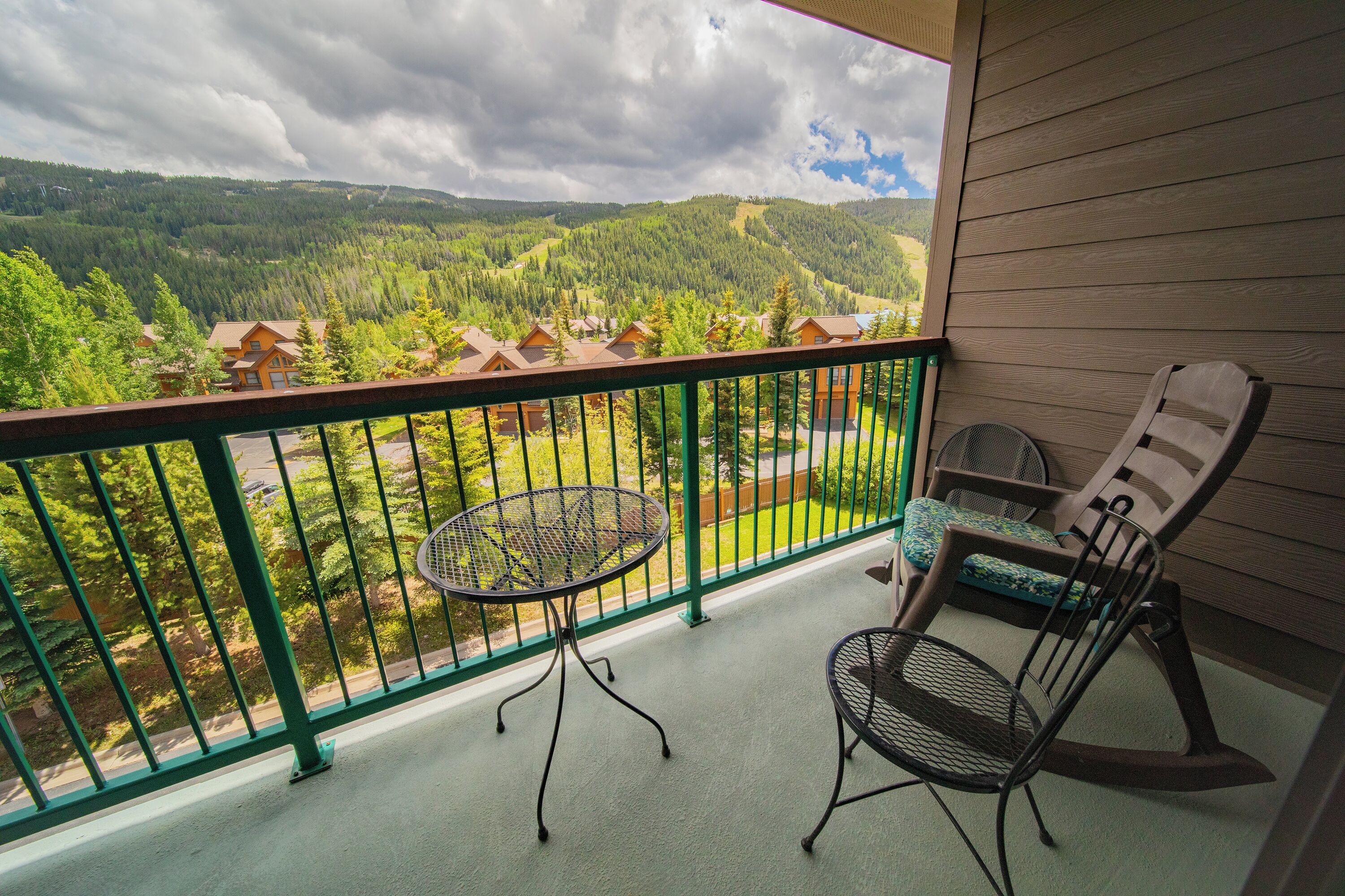 A balcony with a table and chairs, overlooking a forested valley with houses.