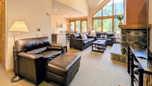A spacious living area with leather sofas, a stone fireplace, and large windows offering a view of the forest.