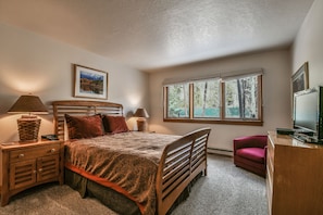 A bedroom with a wooden bed, two bedside lamps, a nightstand, a desk with a chair, a window with a view of trees, and a framed picture on the wall. - Quicksilver 1660 (Keystone)