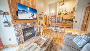 A cozy living room with a stone fireplace, a large TV mounted above it, and a modern kitchen in the background.