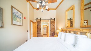 A large bed with white linens, a wooden ceiling, and a chandelier. There are wooden doors leading to other rooms.