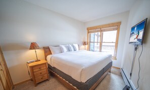 A bedroom with a bed, two bedside tables, a lamp, a window with blinds, and a TV mounted on the wall. - Tenderfoot Lodge 2626 (Keystone)