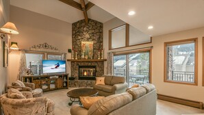 A cozy living room with a stone fireplace, comfortable seating, a flat-screen TV, and large windows offering a view of the outdoors. - Enclave 12 (Keystone)
