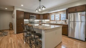 A modern kitchen with wooden cabinets, a stainless steel refrigerator, and a large island with bar stools. - Enclave 12 (Keystone)