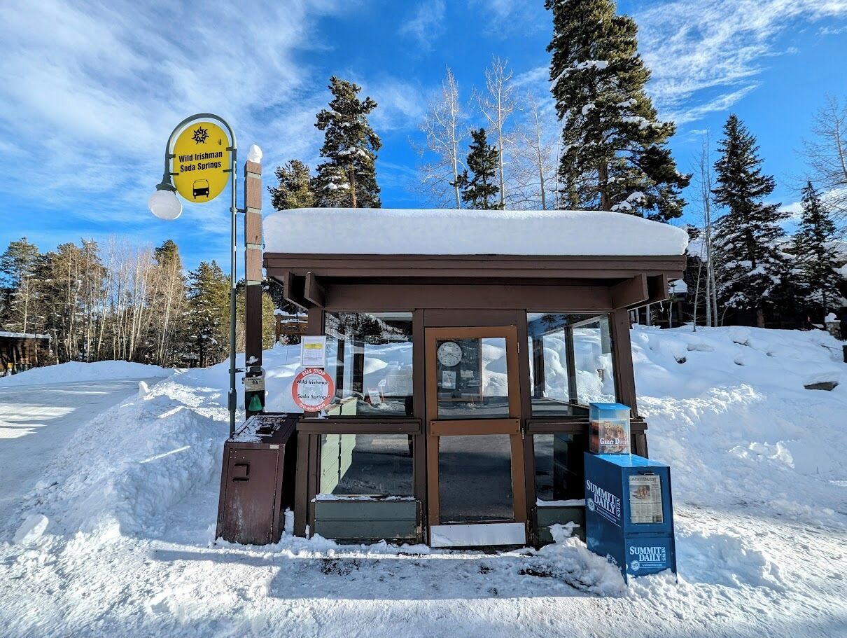 A small, snow-covered building with a glass door, surrounded by snow and trees.