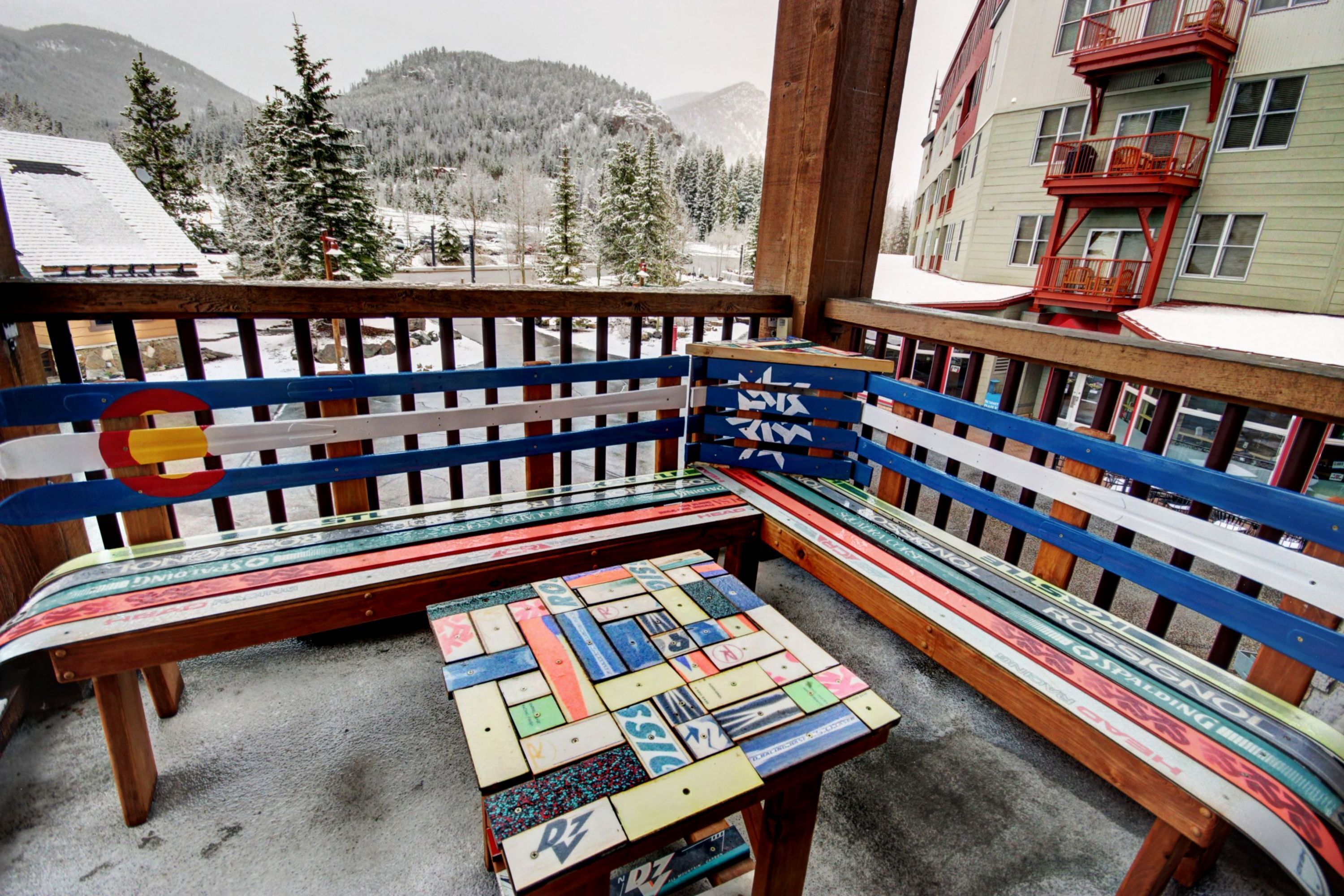 A wooden bench with colorful patterns, a small table, and a view of snow-covered mountains and trees.