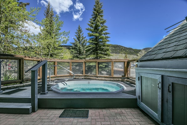 A hot tub with a wooden deck and glass railing, surrounded by trees and a mountain view.