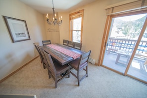 A dining area with a wooden table and chairs, a chandelier, a framed picture, and a view through a glass door to an outdoor deck.