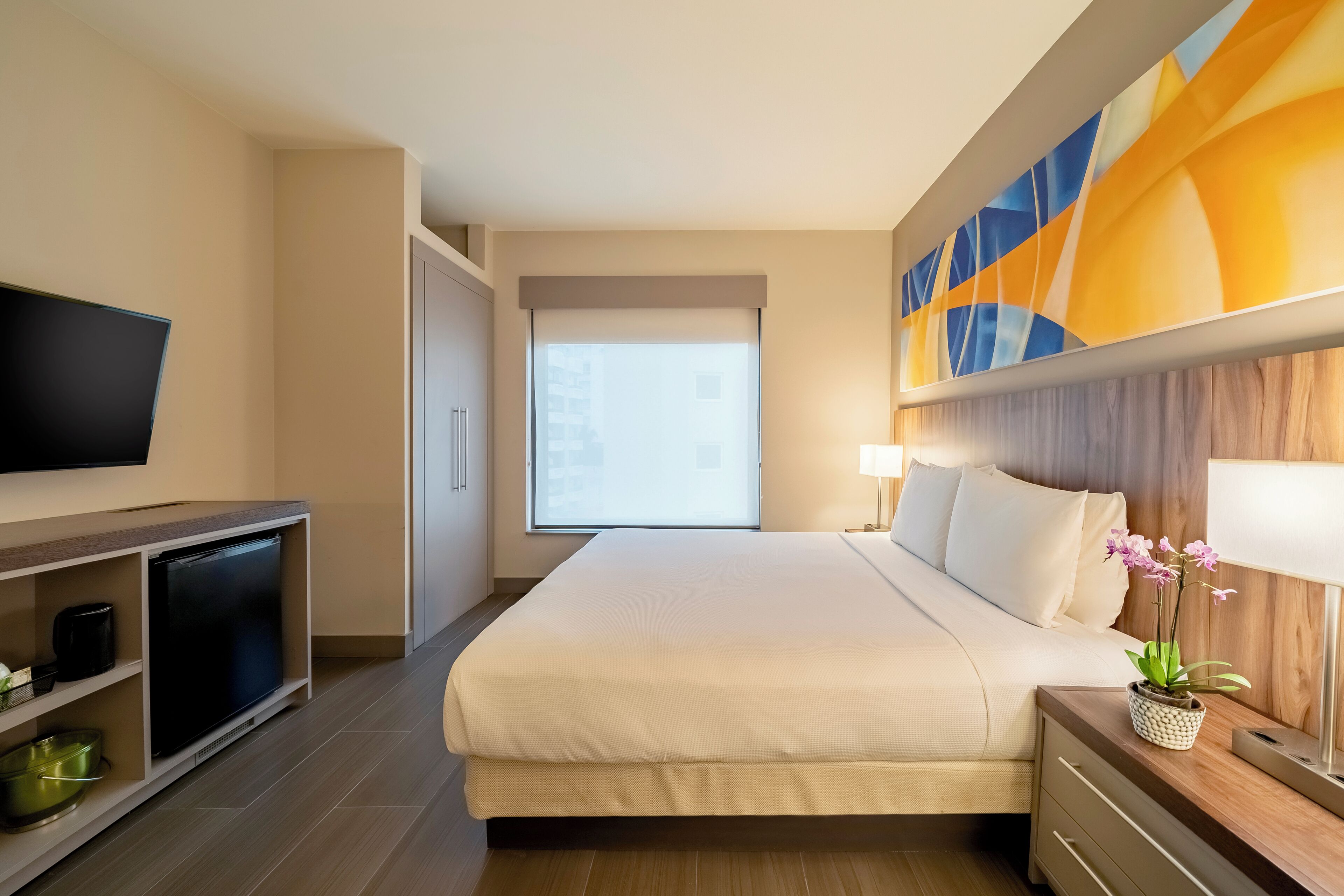 A hotel room with a bed, a television, a desk with a lamp, and a window with a view of buildings.