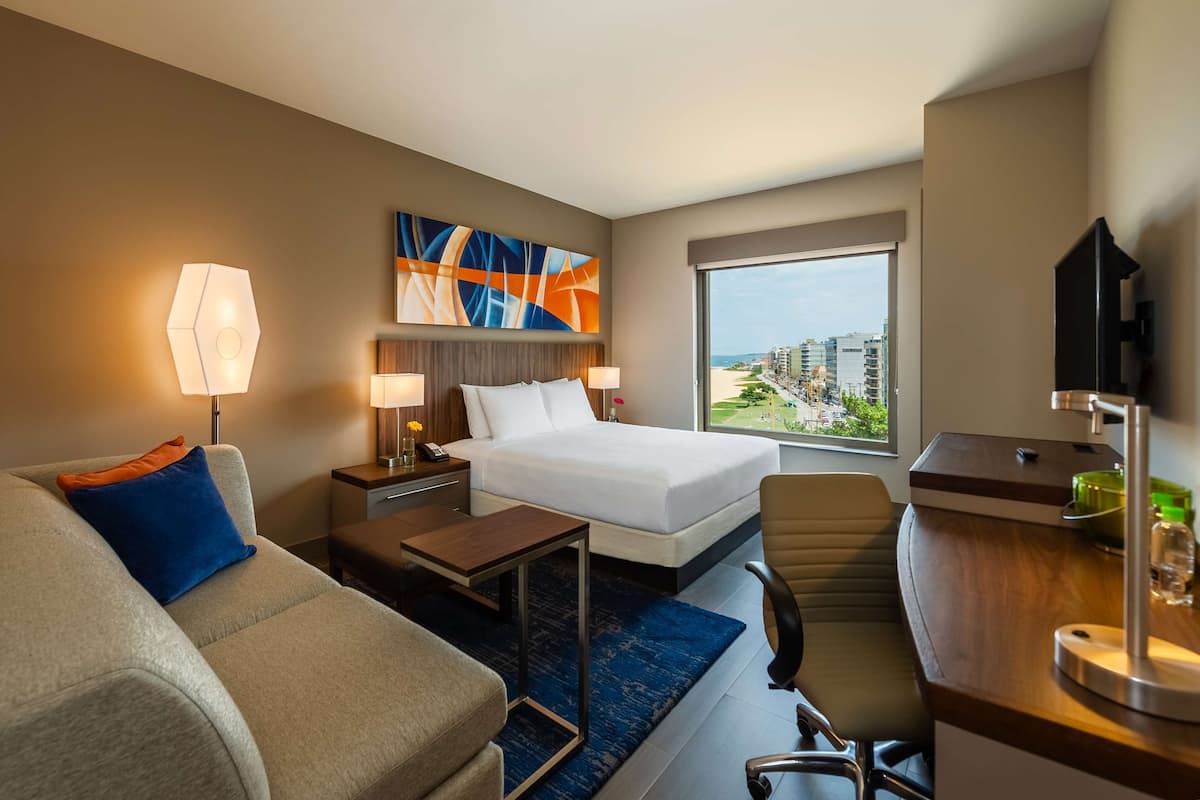 A hotel room with a bed, a desk, a chair, a sofa, and a large window with a city view.