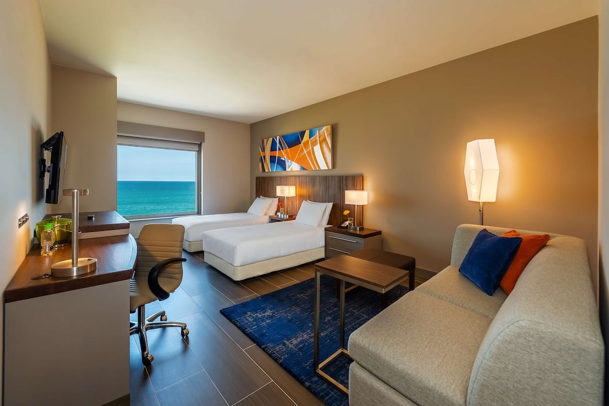 A hotel room with a bed, a desk, a chair, a sofa, and a view of the sea.