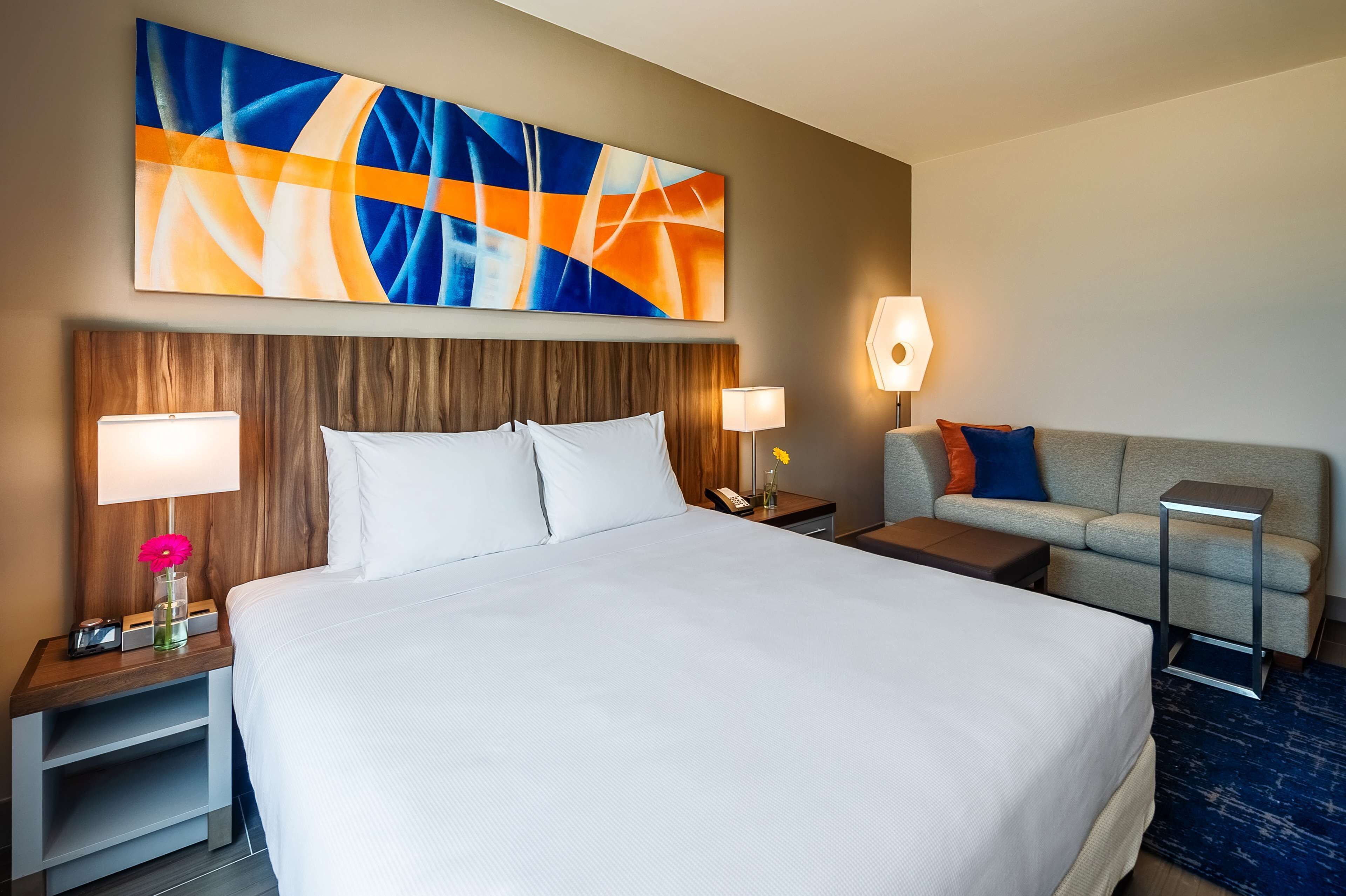 Habitación de hotel con una cama grande, dos mesitas de noche con lámparas, un sofá y un cuadro abstracto colgado en la pared.