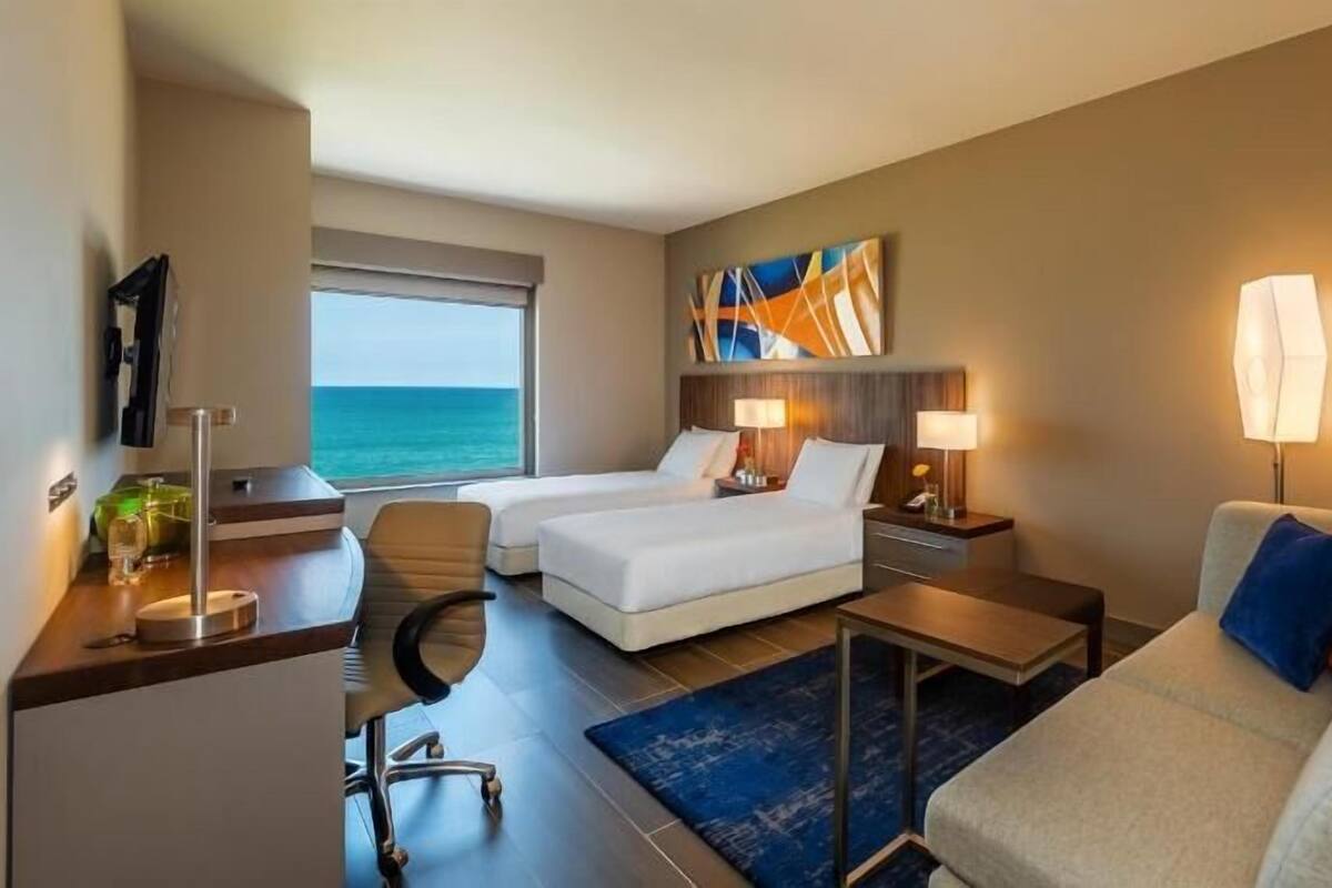 A hotel room with two beds, a desk, a chair, a sofa, and a view of the ocean.