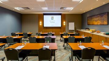 Un salón de conferencias con varias mesas y sillas, una pantalla de proyector mostrando la marca 'HYATT PLACE', y una pizarra.