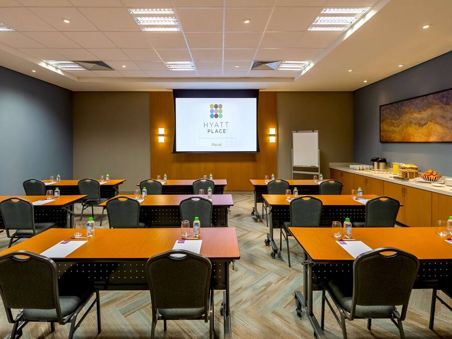 Un salón de conferencias con varias mesas y sillas, una pantalla de proyector mostrando la marca 'HYATT PLACE', y una pizarra.