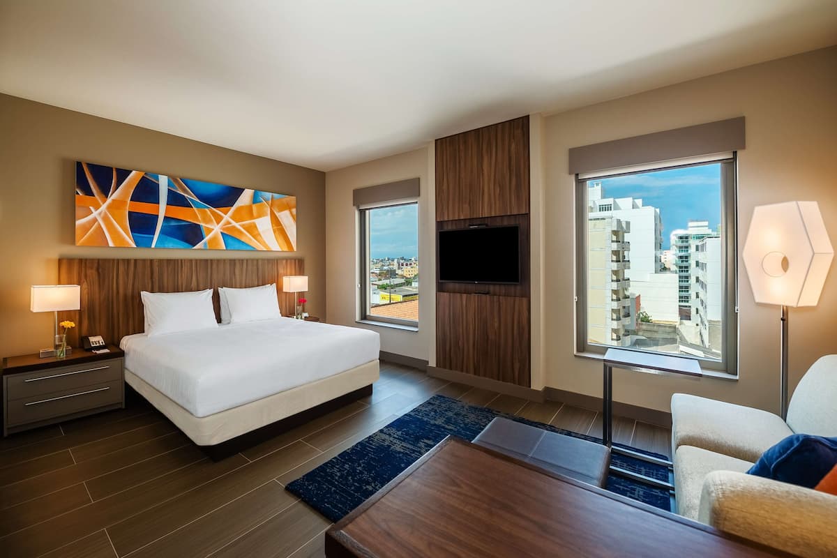 A modern hotel room with a large bed, a flat-screen TV, a desk, and a view of the city.