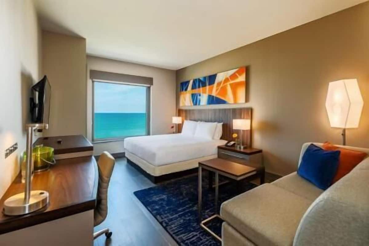 A hotel room with a bed, a desk, a chair, a TV, a window with a view of the sea, and a painting on the wall.