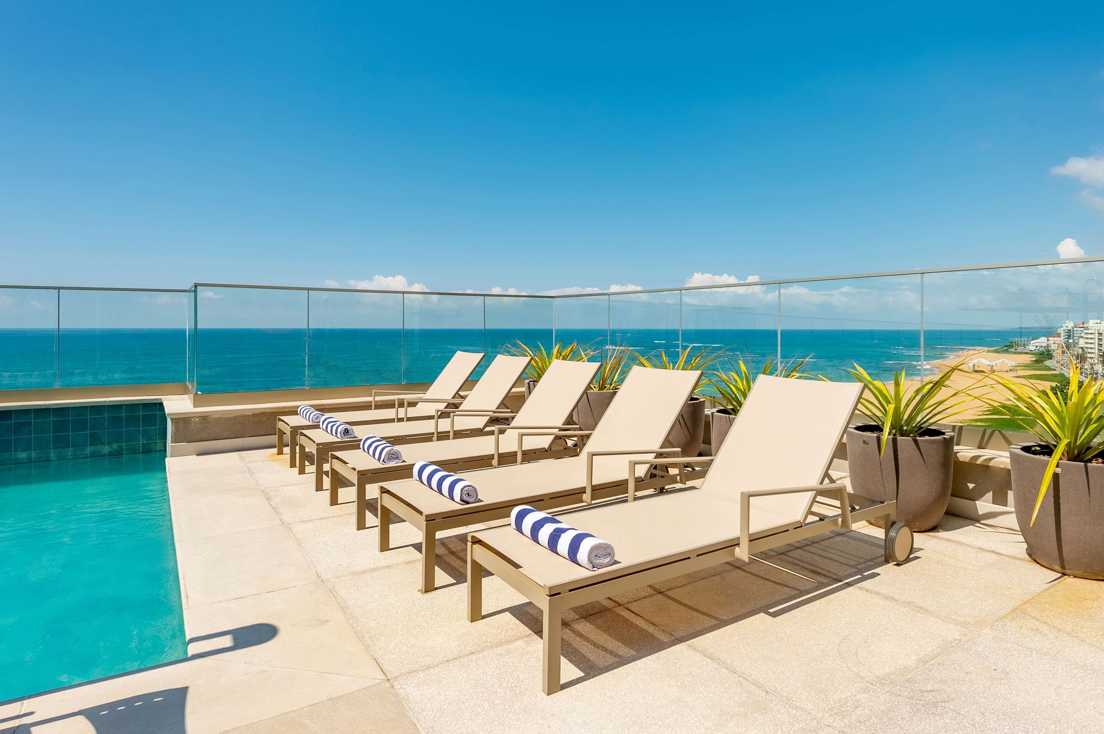 A rooftop pool area with lounge chairs and potted plants, overlooking the ocean.