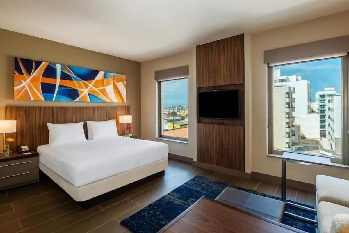 A hotel room with a bed, two bedside tables, a TV, a large window, and a city view.