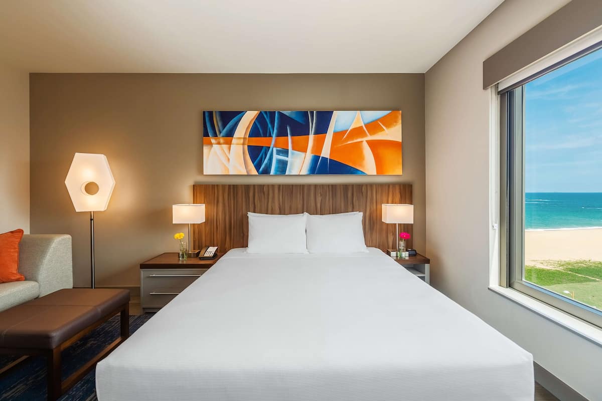 A hotel room with a large bed, two bedside tables with lamps, a nightstand, a sofa, and a painting above the bed.