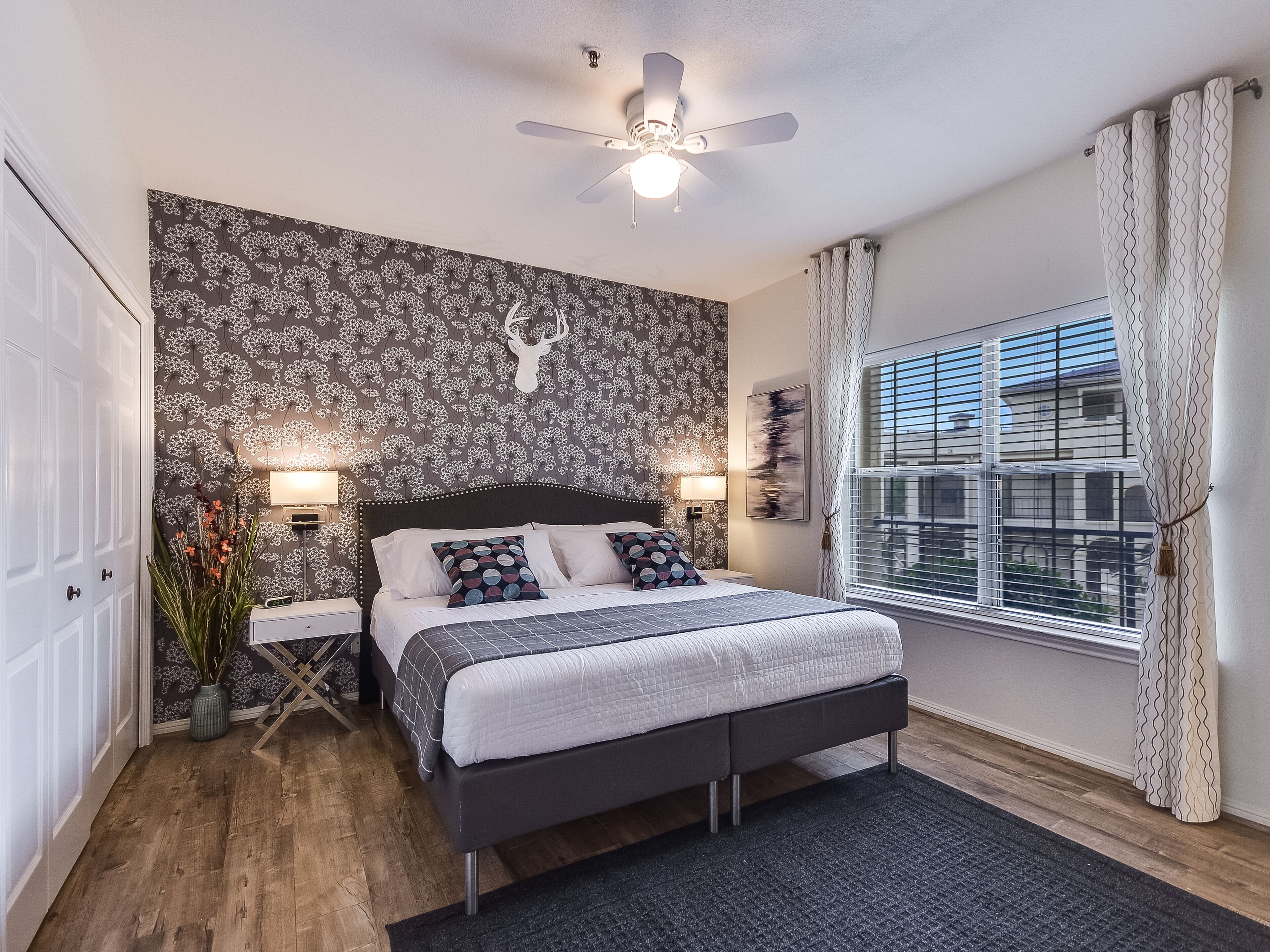 Quarto com cama, mesas de cabeceira, ventilador de teto, janela com cortinas e parede com padrão floral.