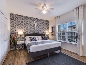 A bedroom with a bed, bedside tables, a ceiling fan, a window with curtains, and a wall with a floral pattern.
