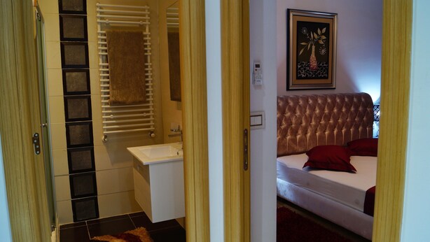 Une salle de bain équipée d’un porte-serviettes, d’un lavabo et d’un miroir. La pièce adjacente contient un lit avec une tête de lit et des oreillers.