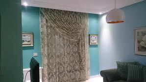 Un salon aux murs de couleur turquoise, avec une grande fenêtre ornée de rideaux, un luminaire au plafond et un canapé agrémenté de coussins.