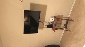 A room with a flat-screen TV mounted on the wall, a wooden table with a computer and a mouse, and a small cabinet. - Wonderful home (4 bdr, 3 bth) cozy! (Cibolo)