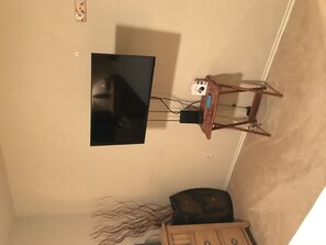 A room with a flat-screen TV mounted on the wall, a wooden table with a computer and a mouse, and a small cabinet.