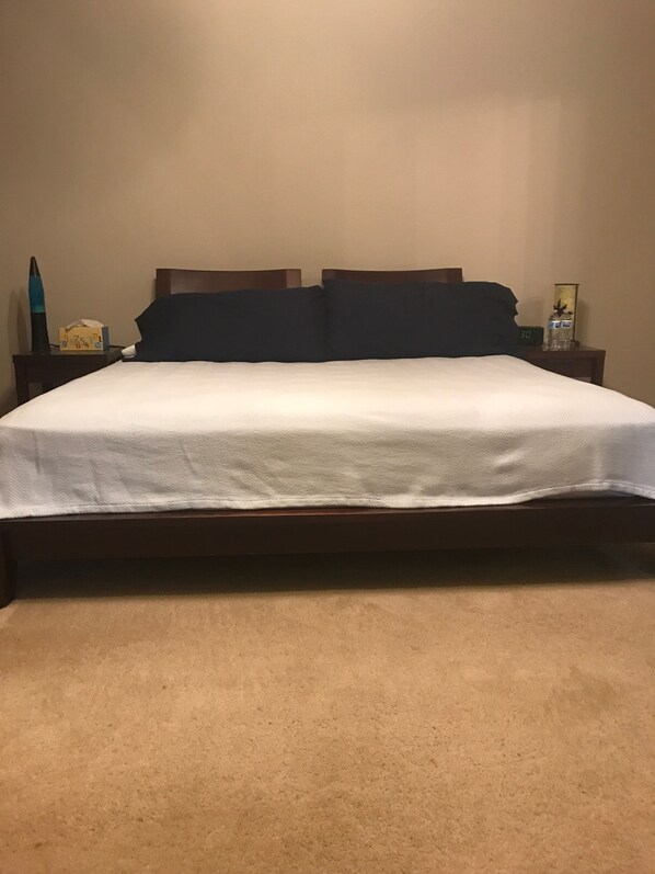 A bed with a white sheet, a wooden headboard, and a dark-colored pillow. There is a nightstand with a clock and a bottle of water. The room has beige carpeting.
