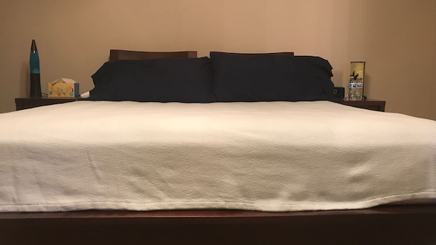 A neatly made bed with a white comforter and dark pillows, a bedside table with a lamp, a tissue box, and a water bottle. - Wonderful home (4 bdr, 3 bth) cozy! (Cibolo)