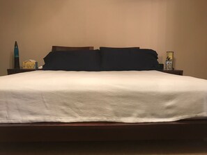 En pænt redt seng med hvid dyne og mørke puder, et sengebord med lampe, tissues og en vandflaske.