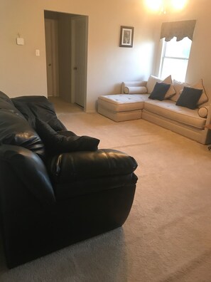 A living room with a black leather sofa, a beige sectional sofa with pillows, a door, and a framed picture on the wall.