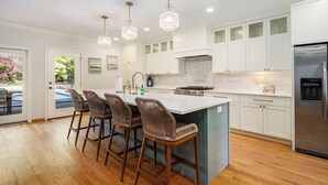 A modern kitchen with white cabinets, a central island with bar stools, pendant lighting, and a stainless steel refrigerator. - Luxury 7BR multifamily on Lake Michigan, Pool & Spa, New Kitchen and Lake Porch! (South Haven)
