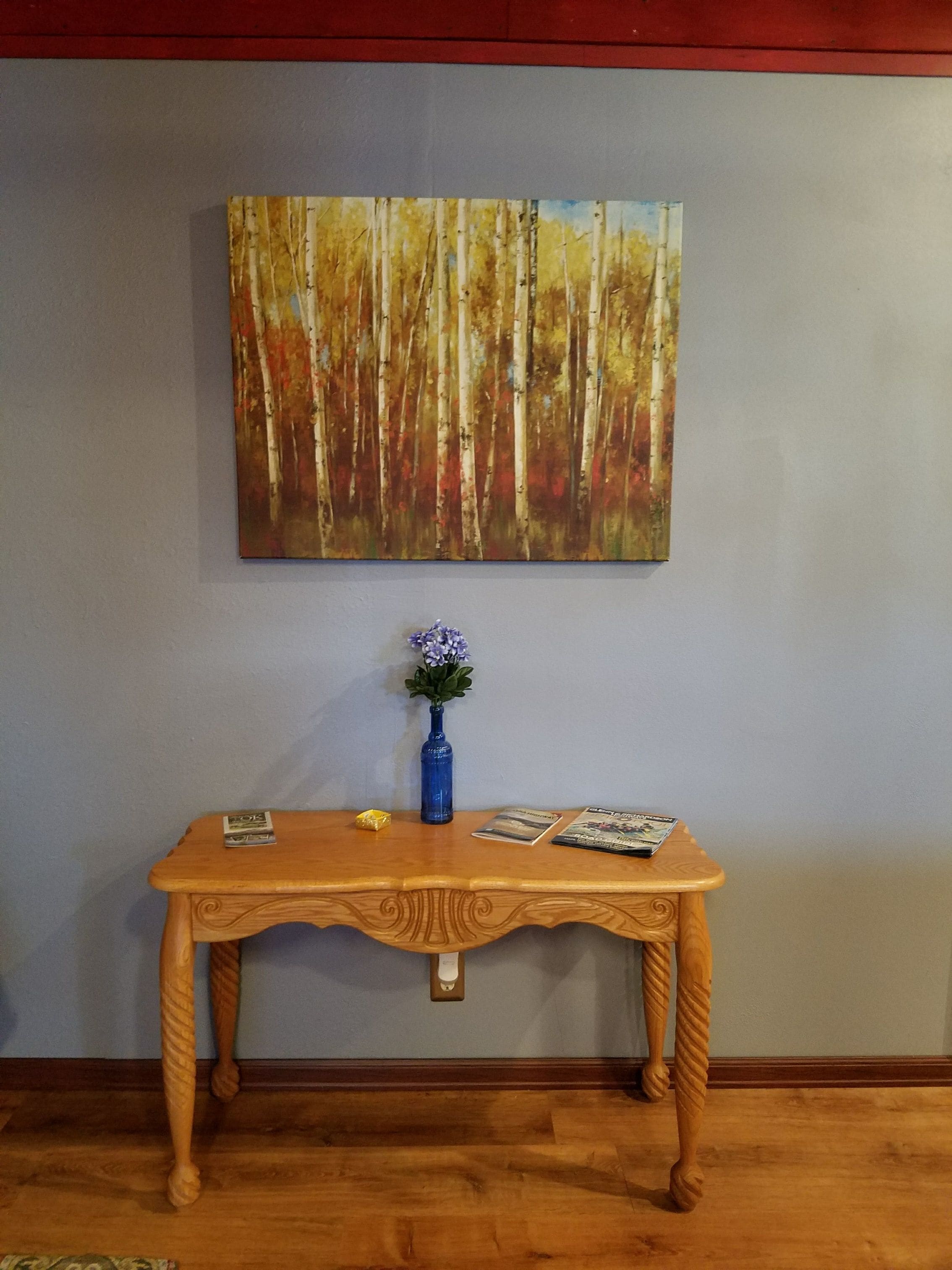 Una mesa de madera con un florero y revistas, frente a una pared con un cuadro de un bosque.
