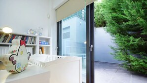 Una habitación moderna con un mostrador de recepción blanco, una estantería con libros y objetos decorativos, y vistas a un árbol verde a través de una puerta de cristal.