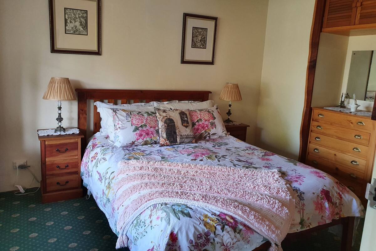 A neatly made bed with floral bedding, two bedside tables with lamps, a dresser with a mirror, and two framed pictures on the wall.