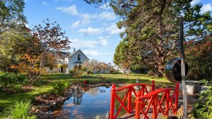 A serene garden with a pond, a red bridge, and a white house in the background. - Belmont Homestead (Swansea)