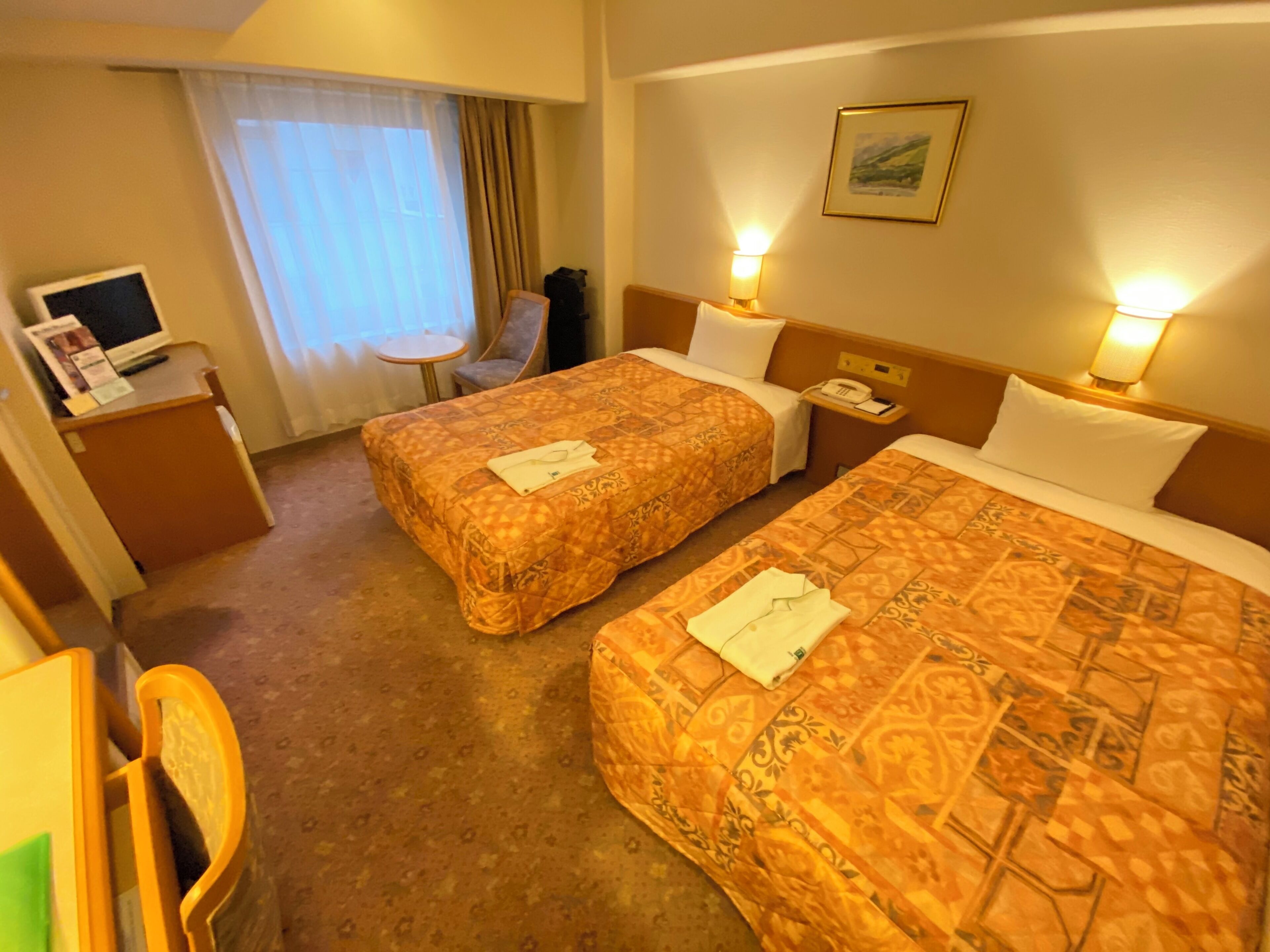 Una habitación de hotel con dos camas, un escritorio con computadora, una silla y una ventana con cortinas.