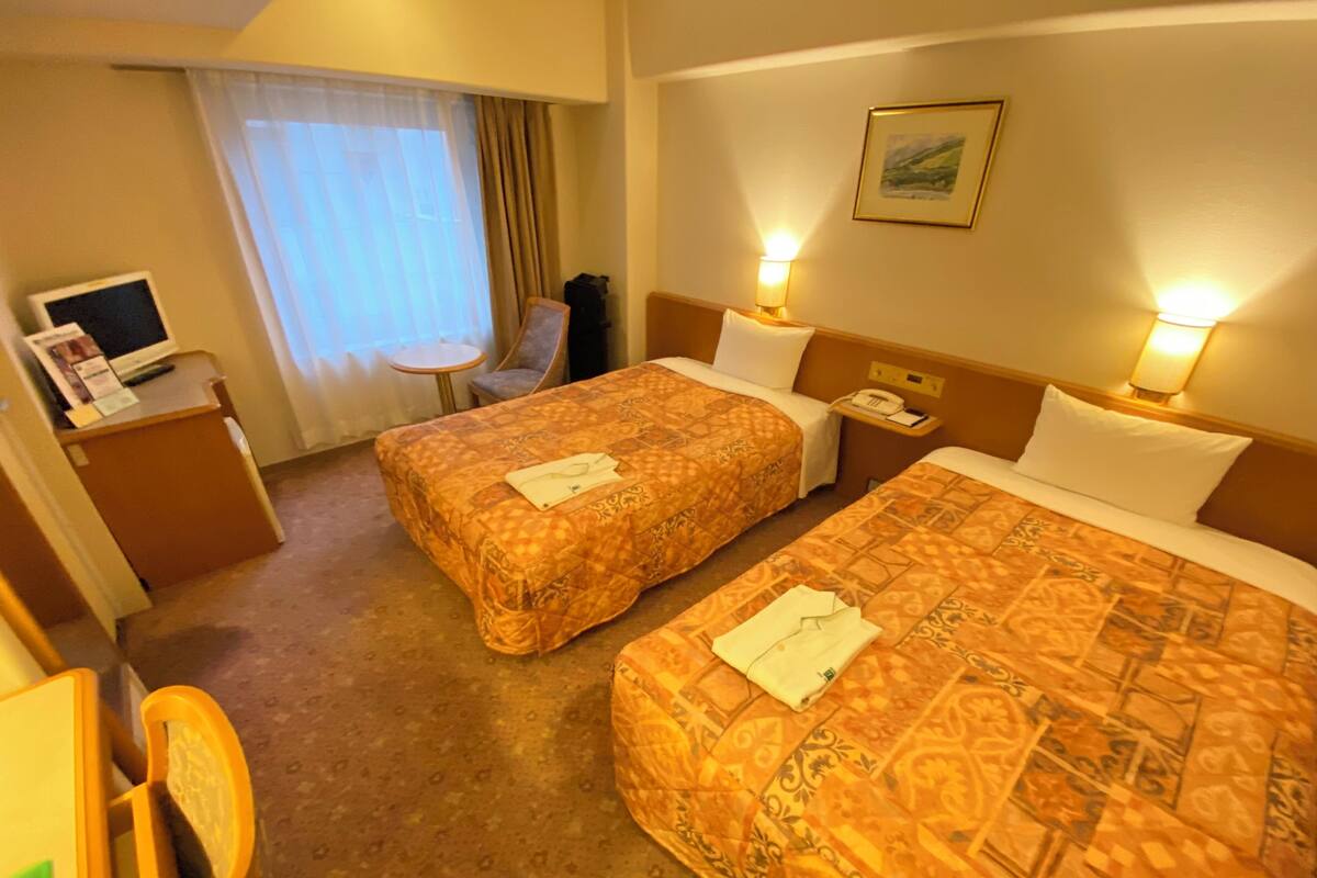 A hotel room with two beds, a desk with a computer, a chair, and a window with curtains.